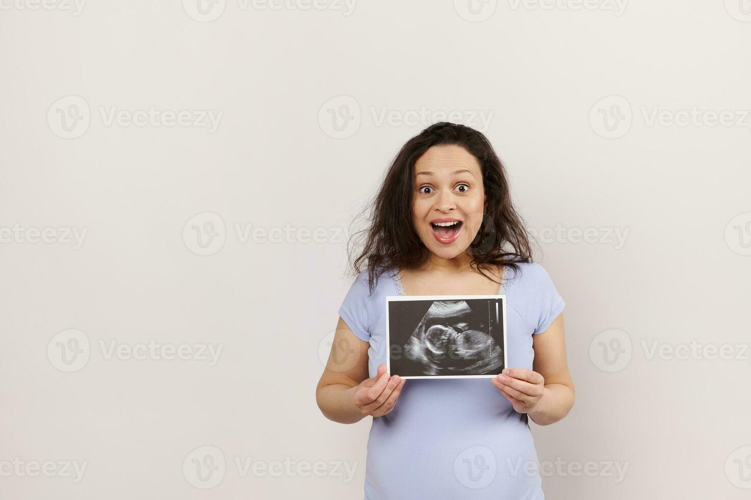 Amazed pregnant woman shows ultrasound scan of newborn baby, expresses positive emotions from pregnancy. White backdrop photo