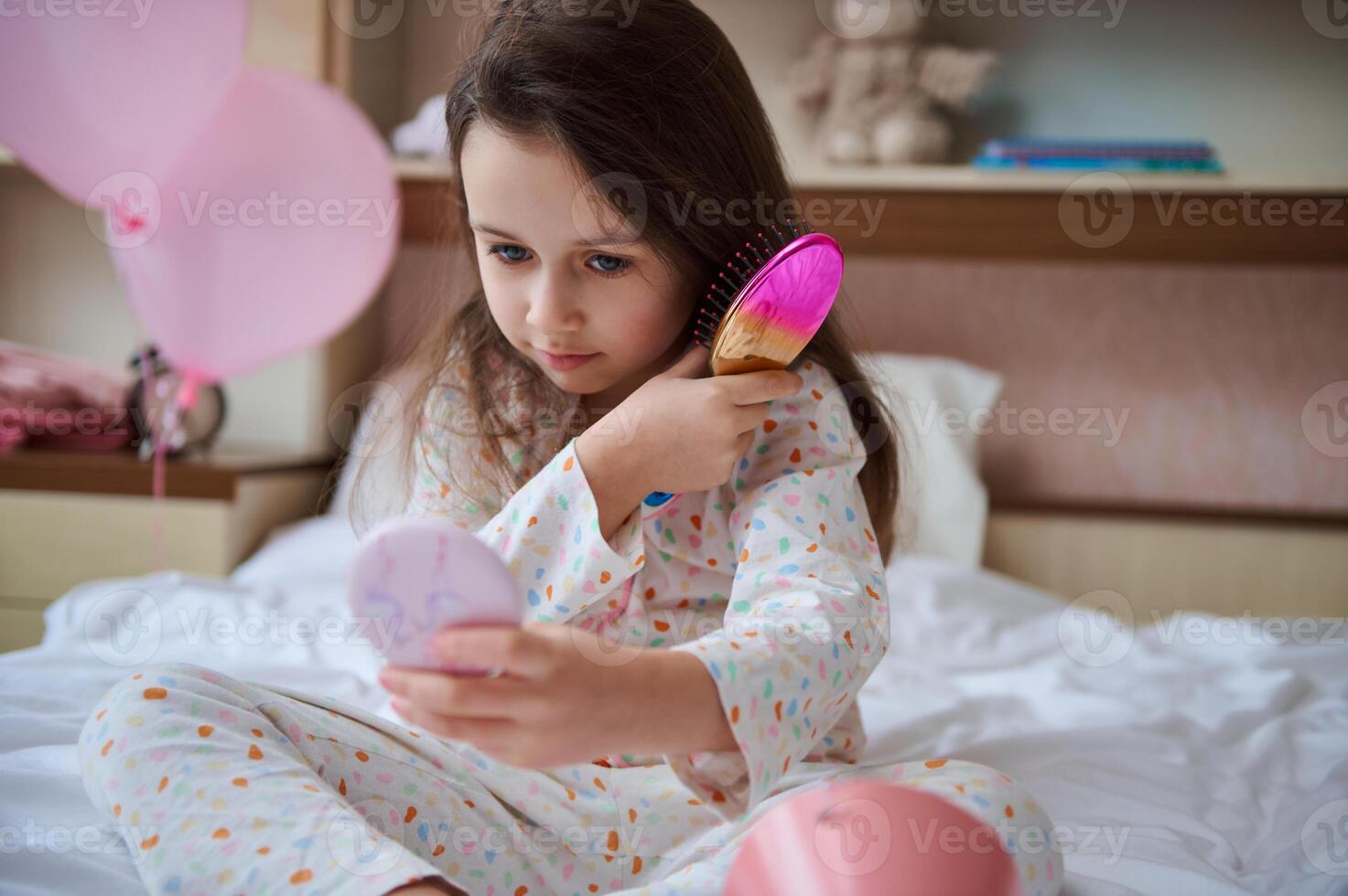 Cute little girl sitting on the bed, looking at her reflection in small cosmetic mirror, combing her beautiful long hair photo