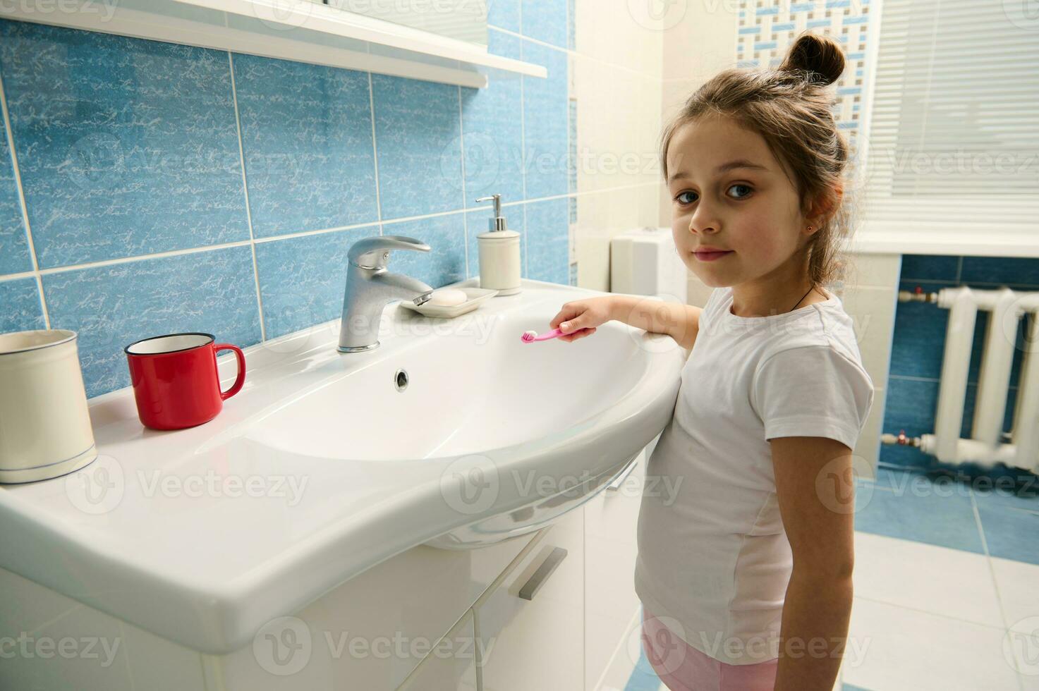 Adorable little girl holding toothbrush, looks at camera standing by sink in the bathroom during morning hygiene routine photo