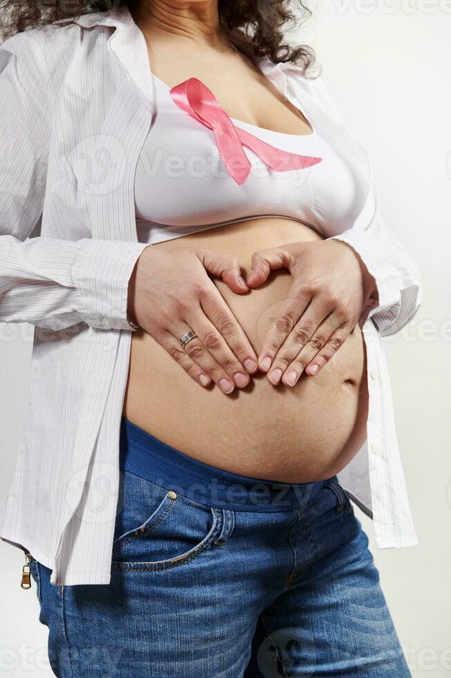 de cerca de embarazada mujer vistiendo rosado cinta, pone manos en su barriga en corazón forma. pecho cáncer conciencia Campaña foto