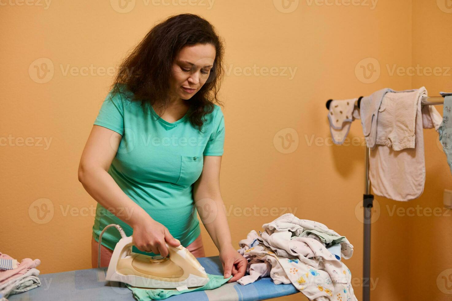 Pregnant woman using steam iron, steaming her newborn bodysuit on ironing board with folded clean crumpled baby clothes photo