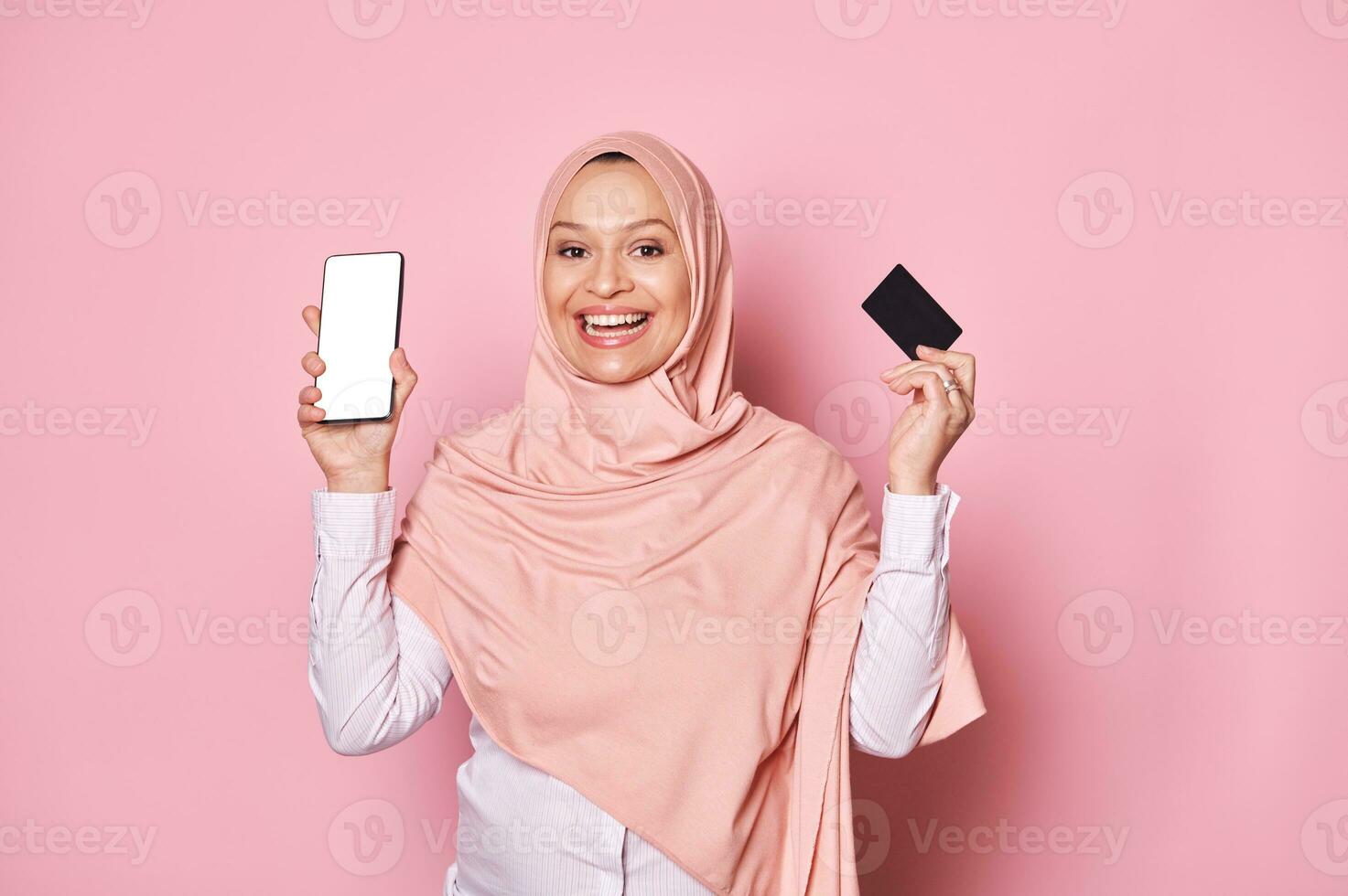 sonriente árabe musulmán mujer en rosado hiyab, participación crédito tarjeta y teléfono inteligente con blanco blanco pantalla, aislado en rosado foto