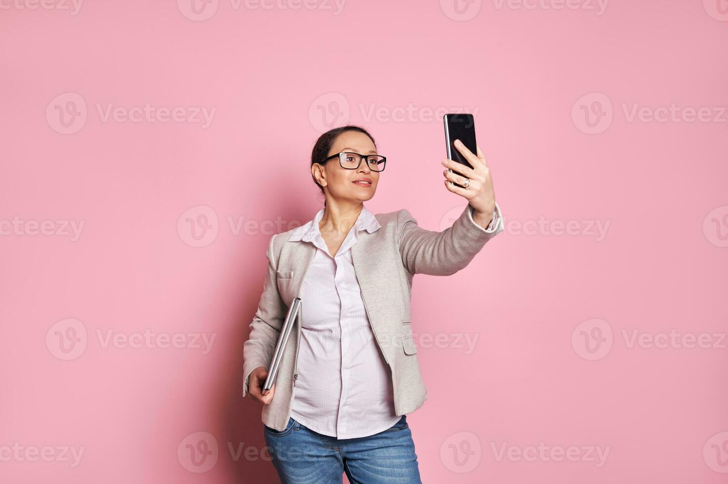 confidente embarazada mujer en casual tener puesto, que lleva computadora portátil, participación móvil teléfono, teniendo en línea conferencia vía vídeo enlace foto