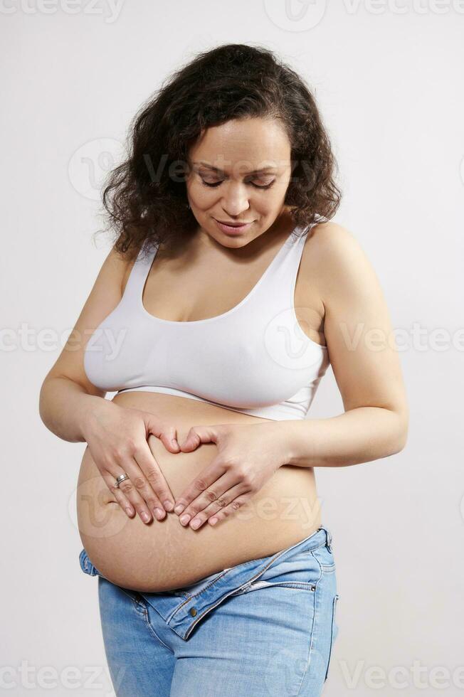 Pregnant woman in white underwear and denim jeans, putting her hands on her belly, making heart shape, isolated on white photo