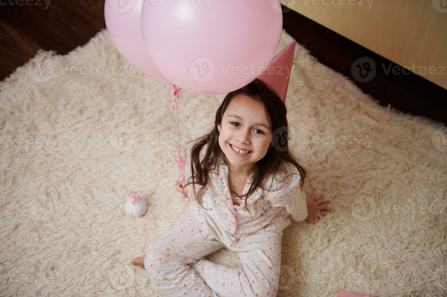 Top view lovely little child, birthday girl in pajamas and pink party hat, sitting on carpet, smiling looking at camera photo