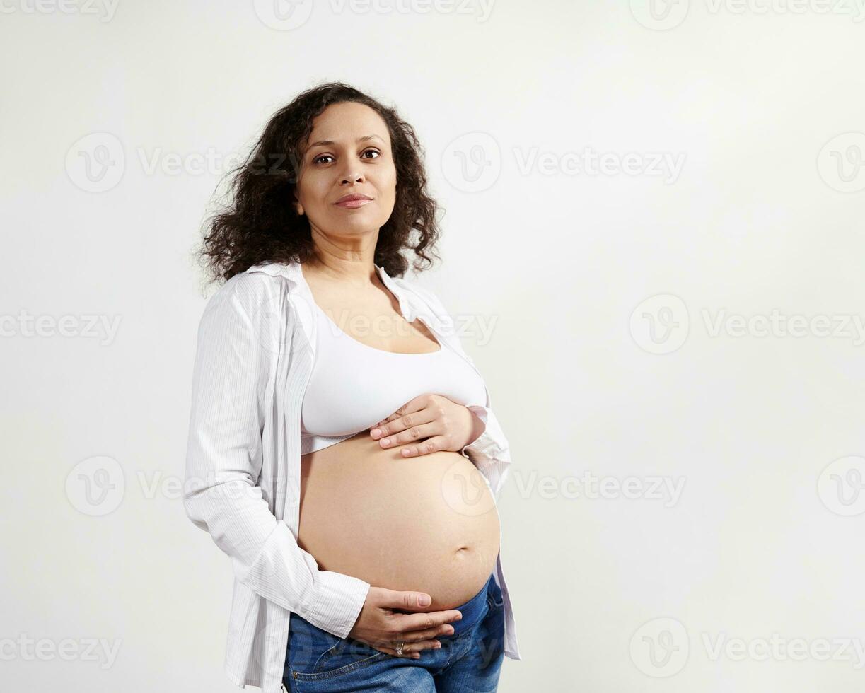 Lovely pregnant ethnic woman dreamily looking away, touching her bare belly, feeling baby kicks. Mothers and Women's Day photo