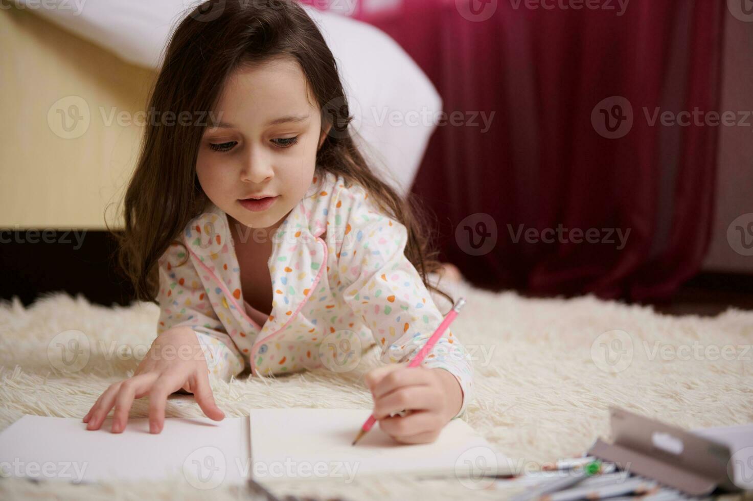 Closeup little girl in pajamas, lying down on carpet in bedroom, concentrated on drawing picture with colorful pencils photo