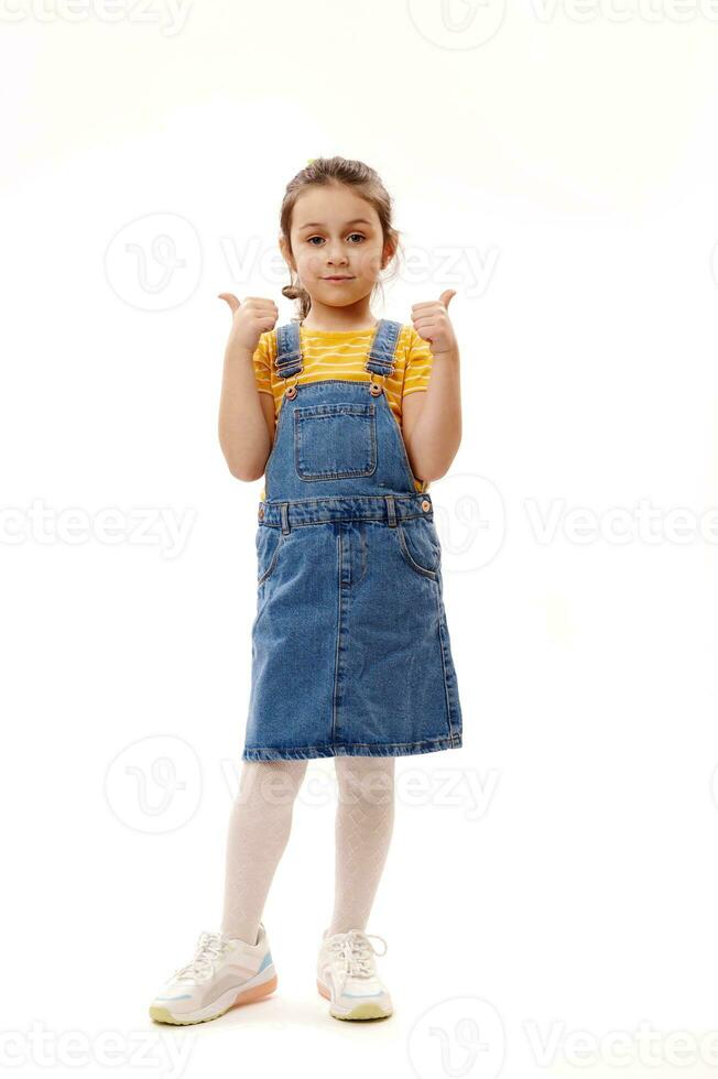 encantador pequeño niña en casual mezclilla ropa, mirando a cámara, gesticulando con pulgares arriba, en pie terminado blanco antecedentes foto