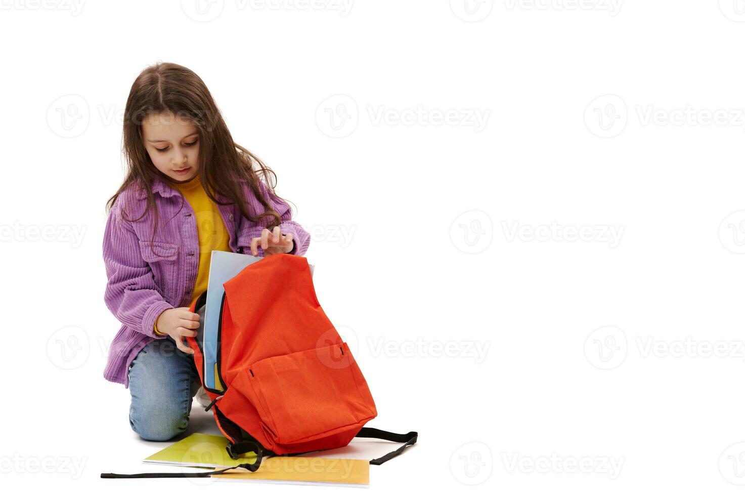 Adorable smart schoolgirl folding textbooks and school supplies into her orange backpack, isolated on a white background photo