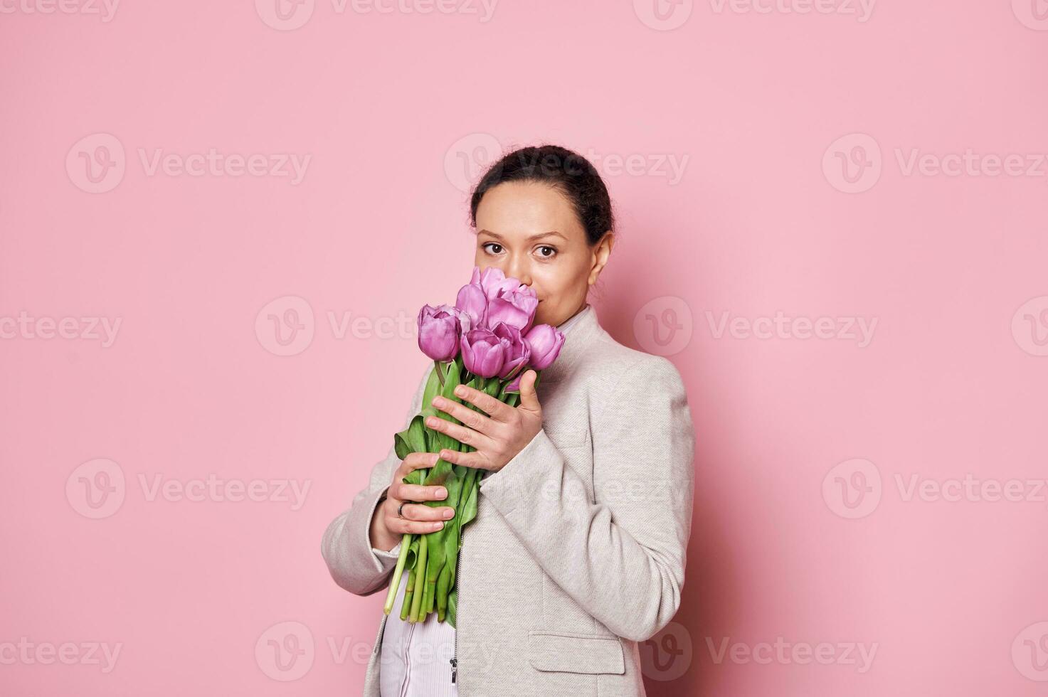 Attractive pregnant woman sniffing a beautiful bouquet of purple tulips, isolated over pink background. Mother's Day photo