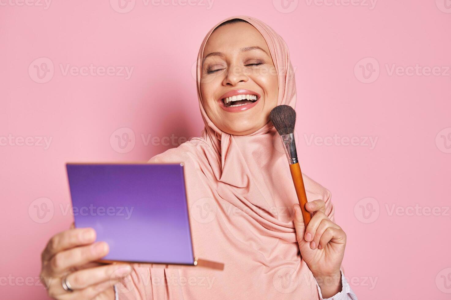 sonriente musulmán mujer en rosado hiyab, aplicando Fundación polvo o sonrojo con maquillaje cepillo en su mejilla, aislado en rosado foto