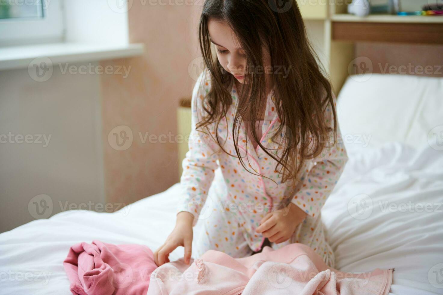 pequeño niña en pijama, plegable su cama y limpia el habitación después despertar arriba en el Mañana. enseñando niños a orden foto