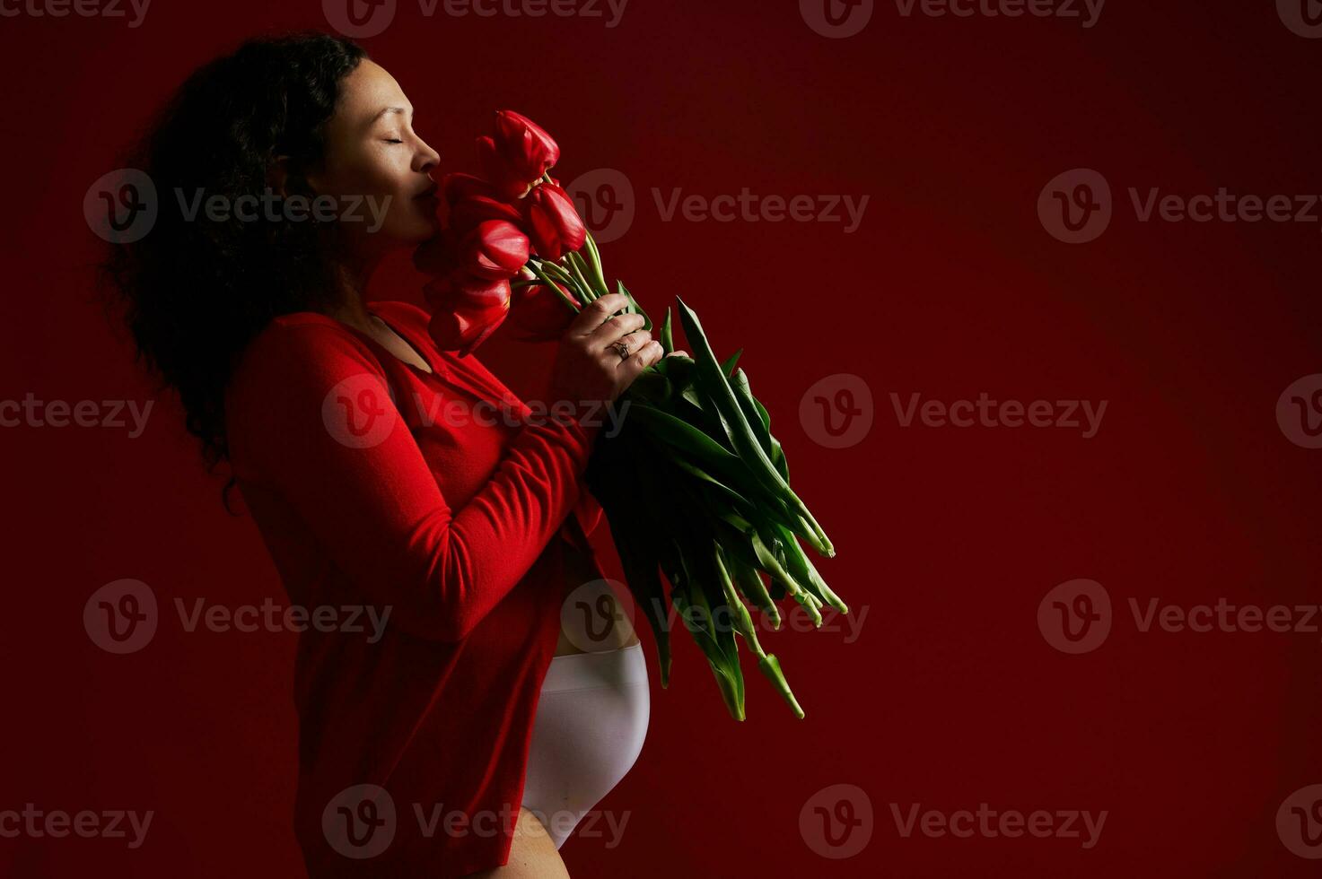 Side portrait on red background of beautiful pregnant woman sniffing a bouquet of red tulips. Happy Mother's Day concept photo