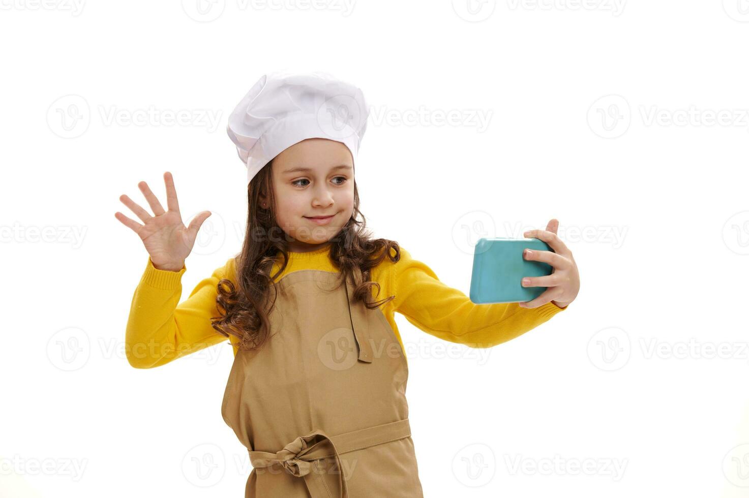 Cute little girl wearing white chef's hat and kitchen apron, taling on mobile phone via video link, isolated background photo