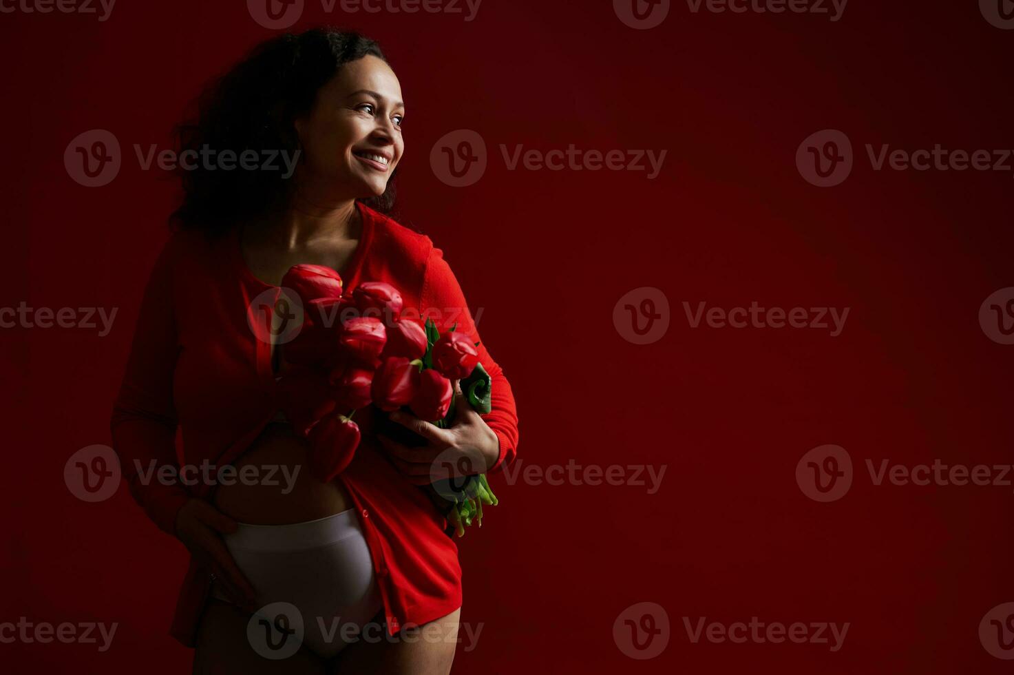 Happy adult pregnant woman caressing her belly, smiling cheerfully looking aside, posing with a bouquet of red tulips photo