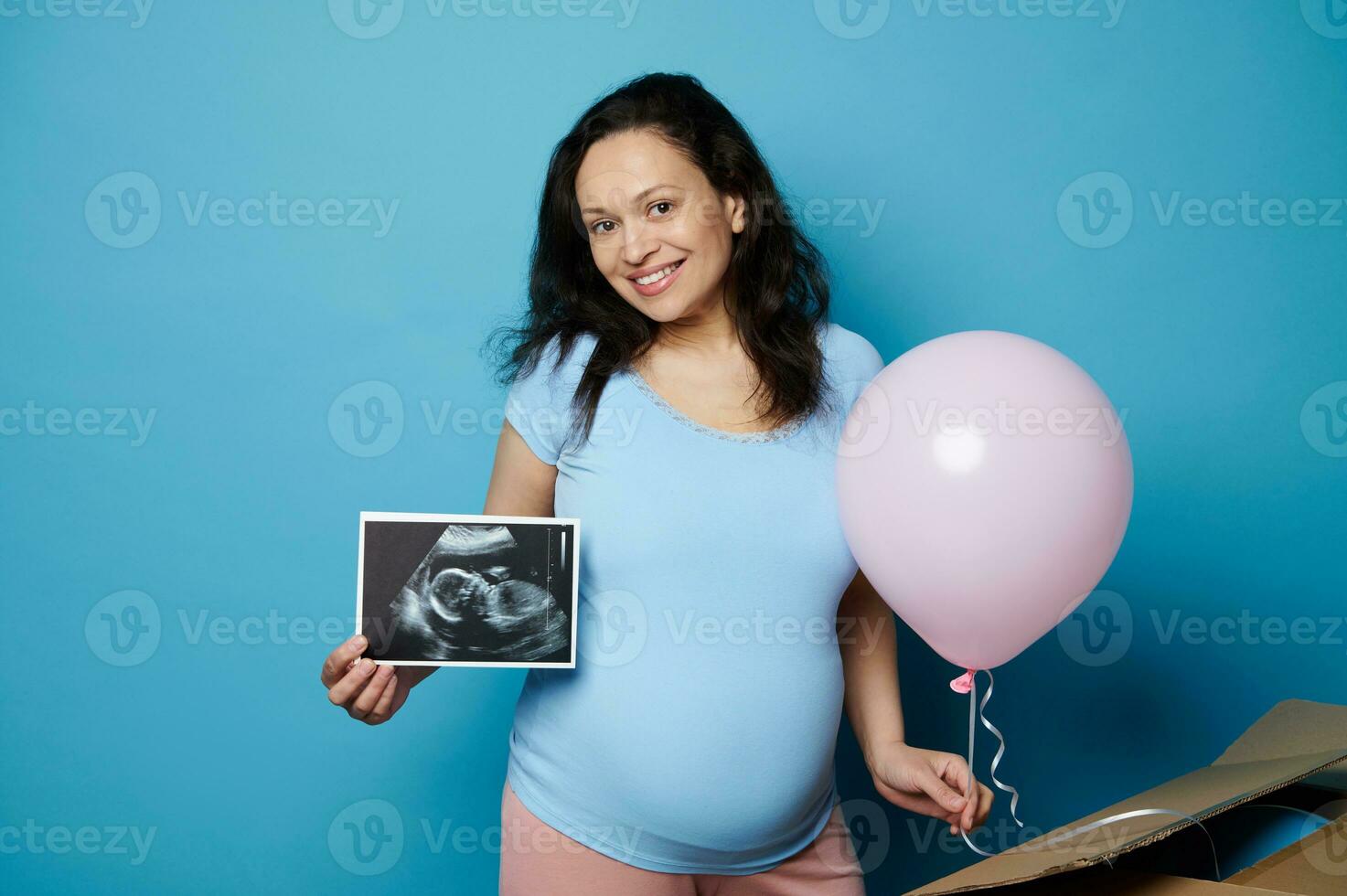 Ethnic pregnant woman with ultrasound scan of her newborn baby, smiles at camera, holds a pink balloon, isolated on blue photo