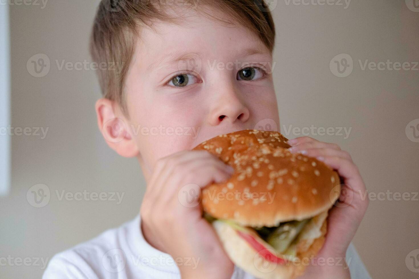 de cerca retrato de un pequeño chico comiendo un enorme hamburguesa, en un blanco antecedentes foto