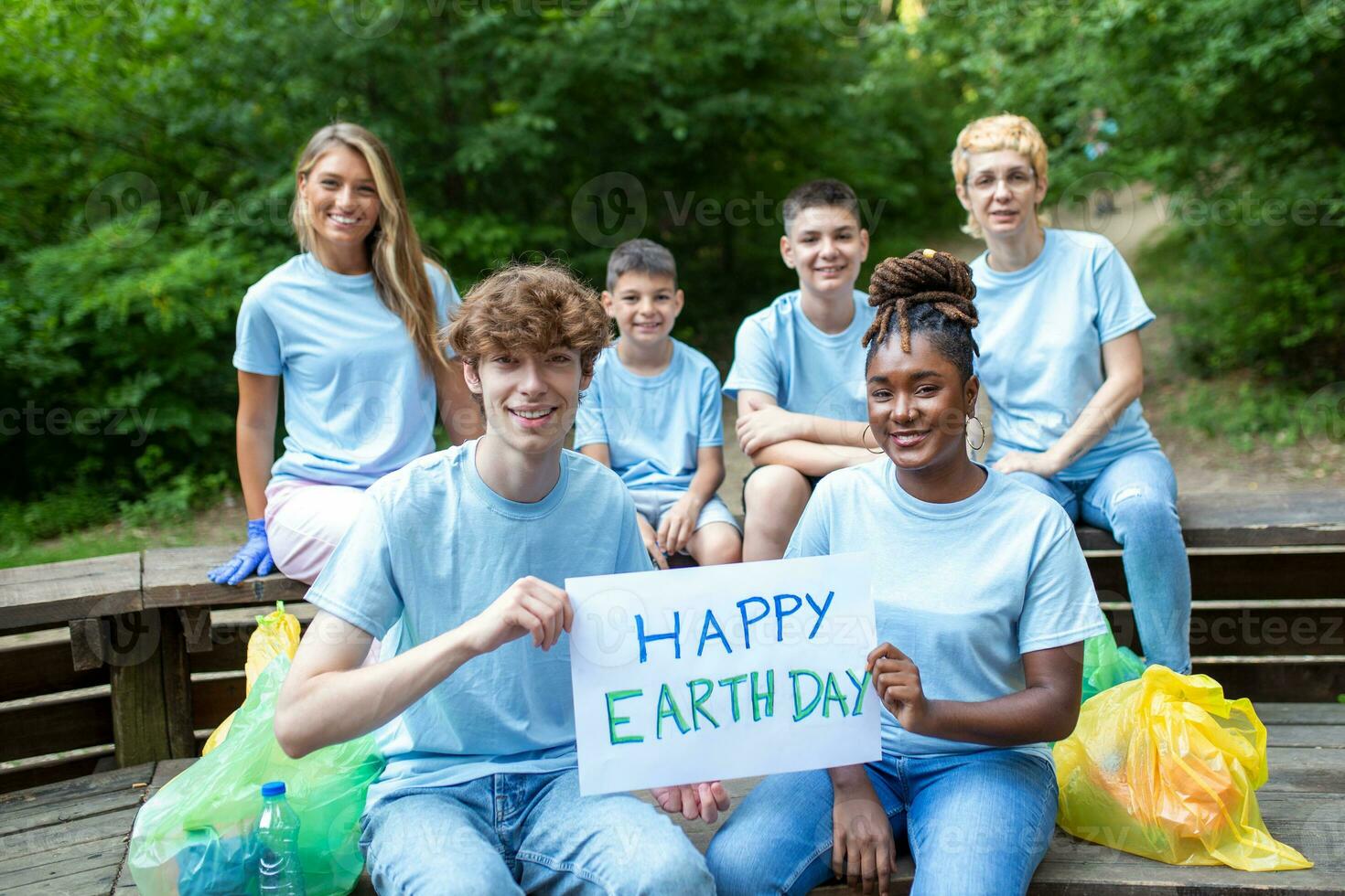 Happy volunteers holding placard with 'happy Earth day' message. Volunteering, charity, cleaning, people and ecology concept - group of happy volunteers with garbage bags cleaning area in park photo