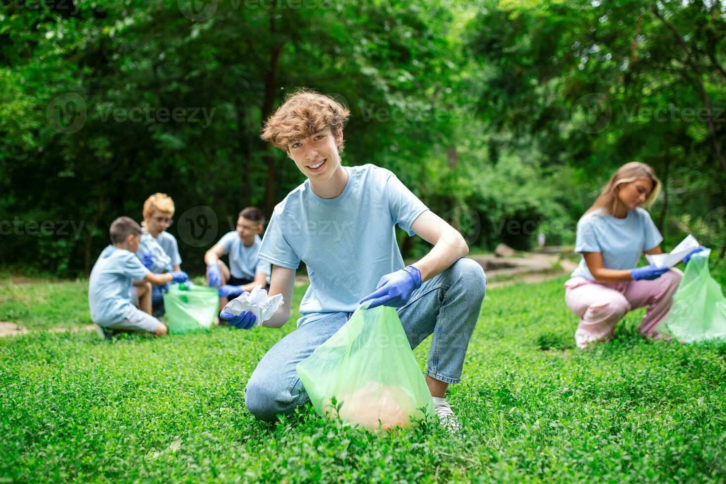 Happy community service people cleaning up local park. They have a trash bag and are filling is with trash they have found in the park. Their are two knealing down and others behind them photo