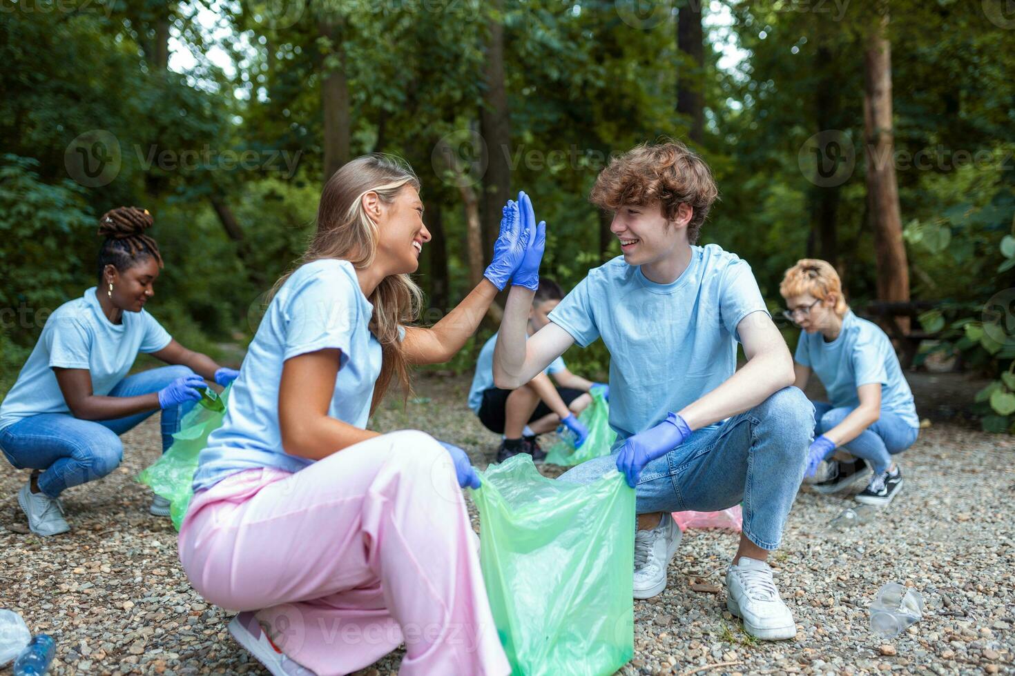 Happy community service people cleaning up local park. They have a trash bag and are filling is with trash they have found in the park. Their are two knealing down and two others behind them photo