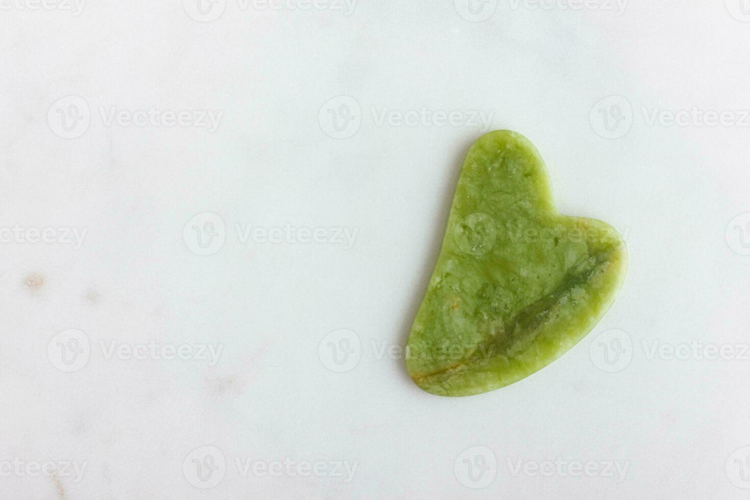 verde jade gua sha masajeador para cara en blanco mármol. foto