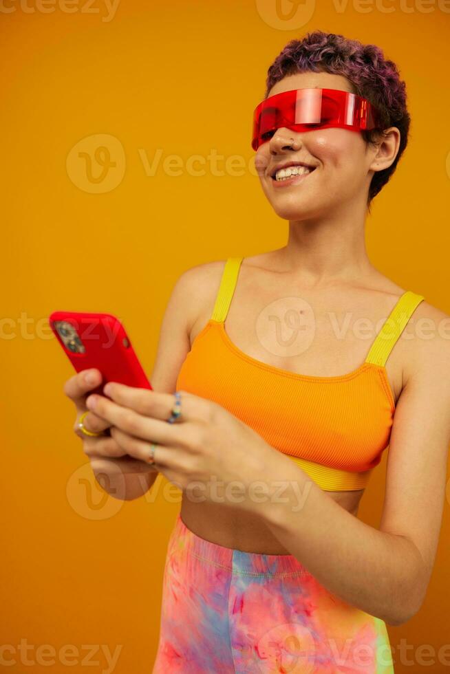 Woman blogger smiling in unusual red millennial glasses holding her phone and looking at the screen against an orange background in a stylish yellow top photo
