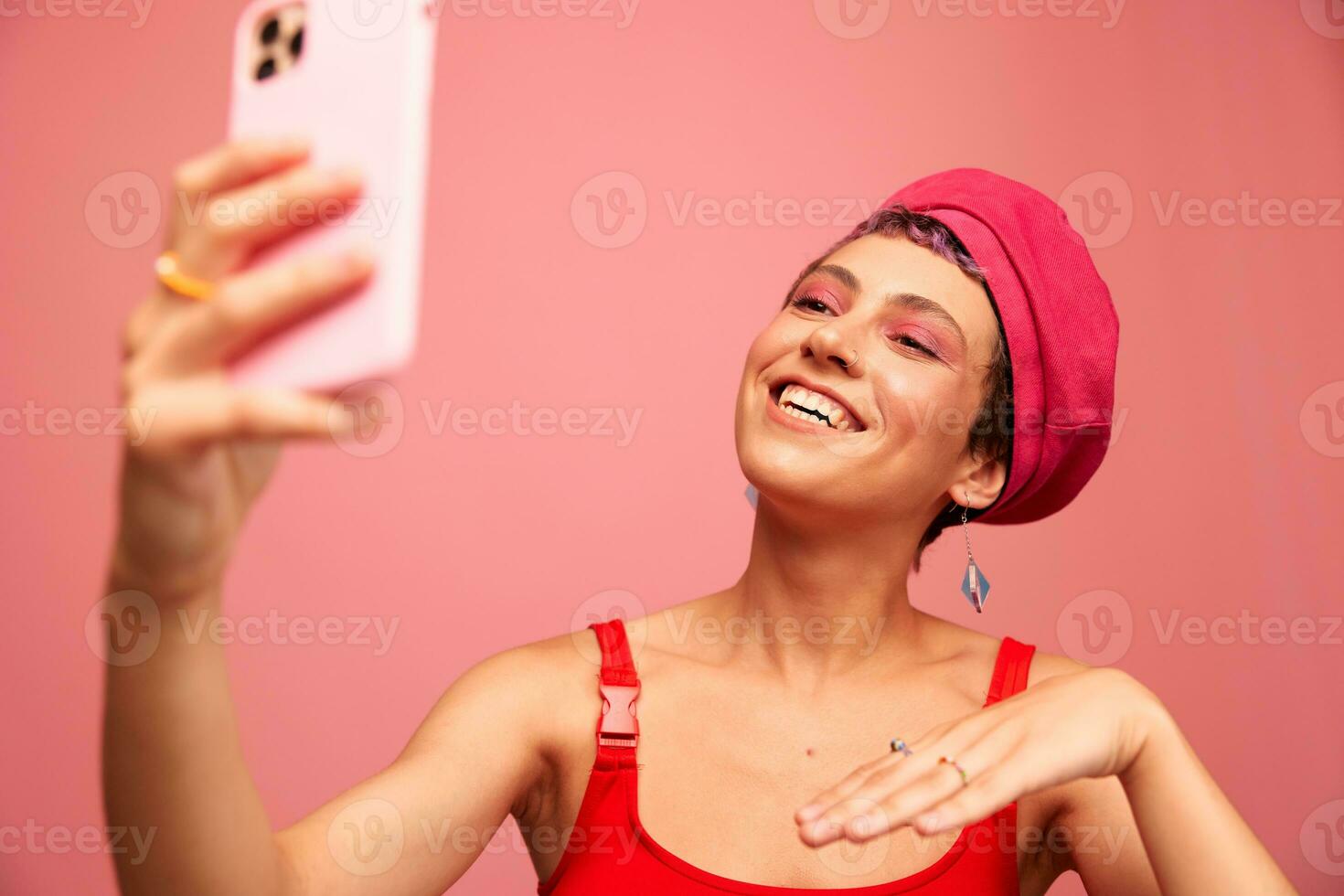 A young woman blogger with colored pink hair and a short haircut takes a picture of herself on the phone and broadcasts a smile in stylish clothes and a hat on a pink background monochrome style photo
