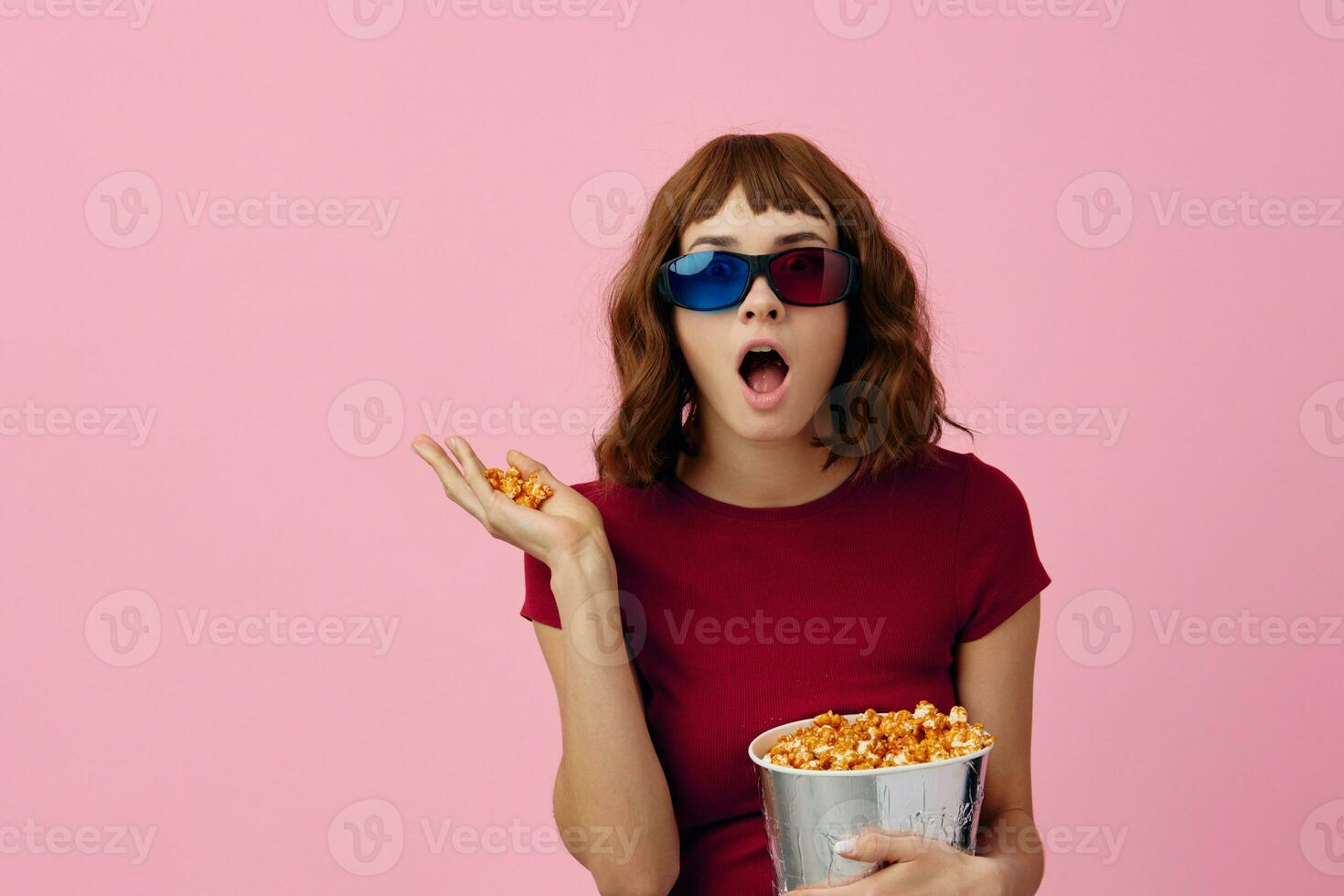 Overjoyed happy cute redhead lady in red t-shirt 3D glasses with popcorn watch movie posing isolated on over pink studio background. Copy space Banner. Fashion Cinema concept. Entertainment offer photo