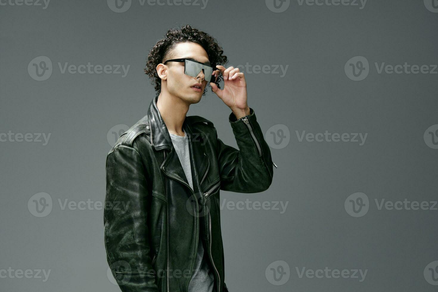 Stylish tanned curly man leather jacket trendy mirror sunglasses posing isolated on over gray studio background. Cool fashion offer. Huge Seasonal Sale New Collection concept. Copy space for ad photo