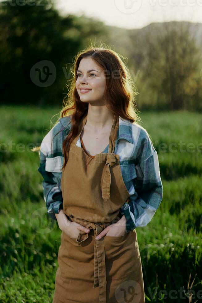 un mujer jardinero en un delantal soportes en un campo de verde césped al aire libre, sonriente en un verano día dentro un puesta de sol foto