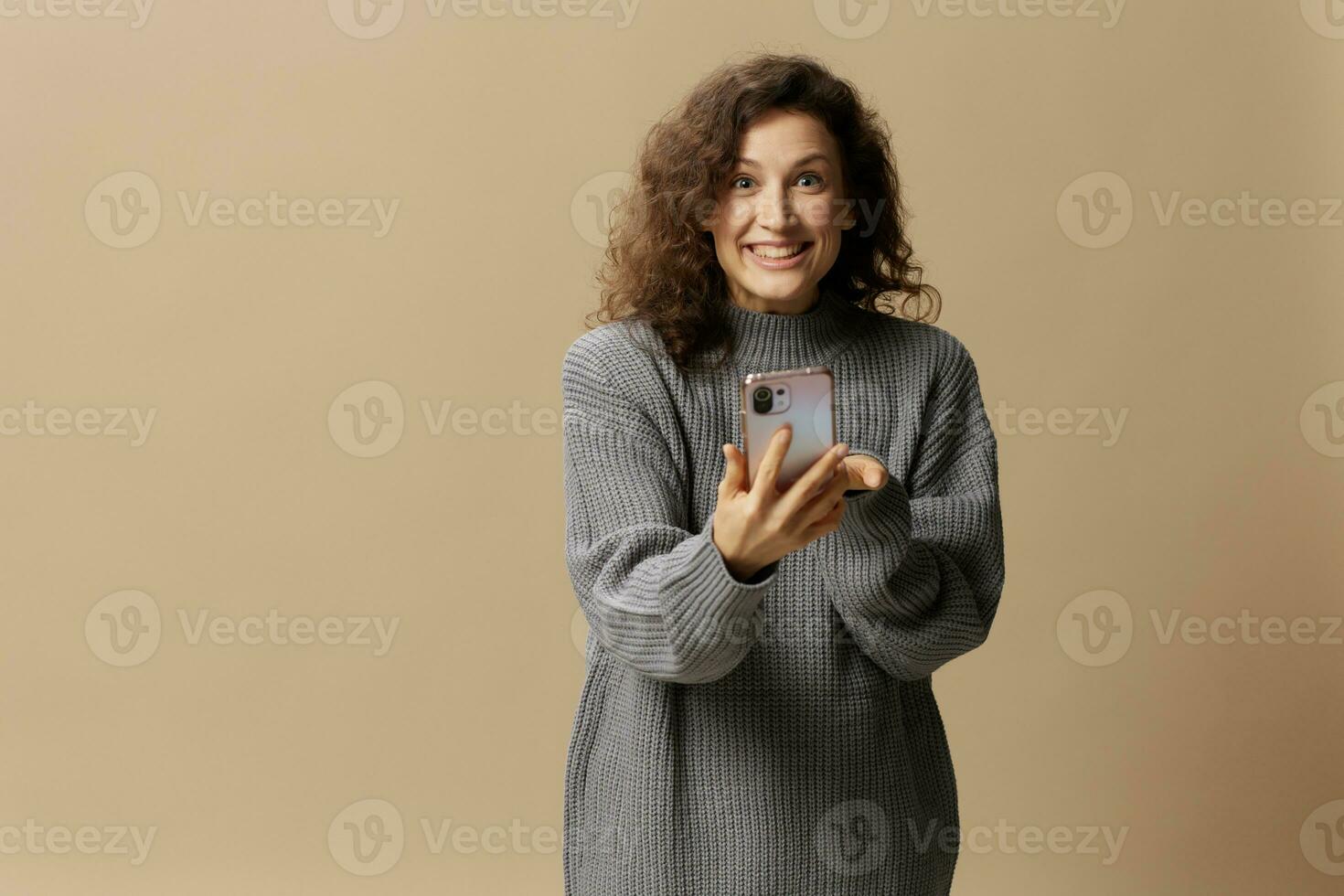 Overjoyed lucky curly cute lady in gray casual sweater get cool news from message posing isolated on over beige pastel background. Social media, network, distance communication concept. Copy space photo