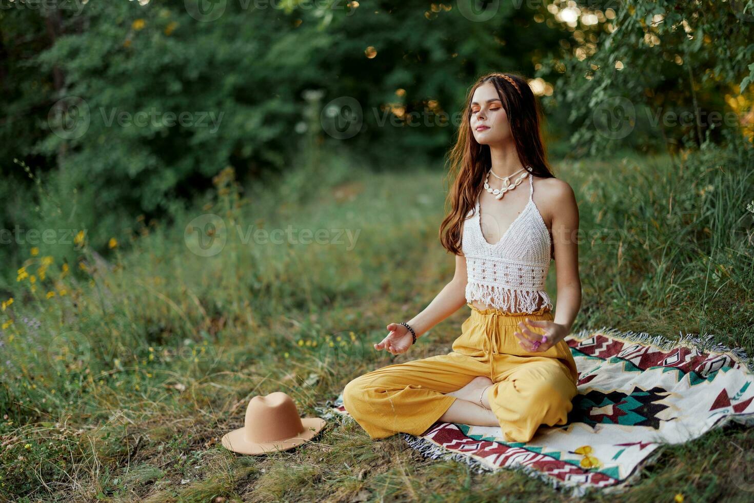 A young hippie woman meditates in nature in the park, sitting in a lotus position on her colorful plaid and enjoying harmony with the world in eco-clothing photo