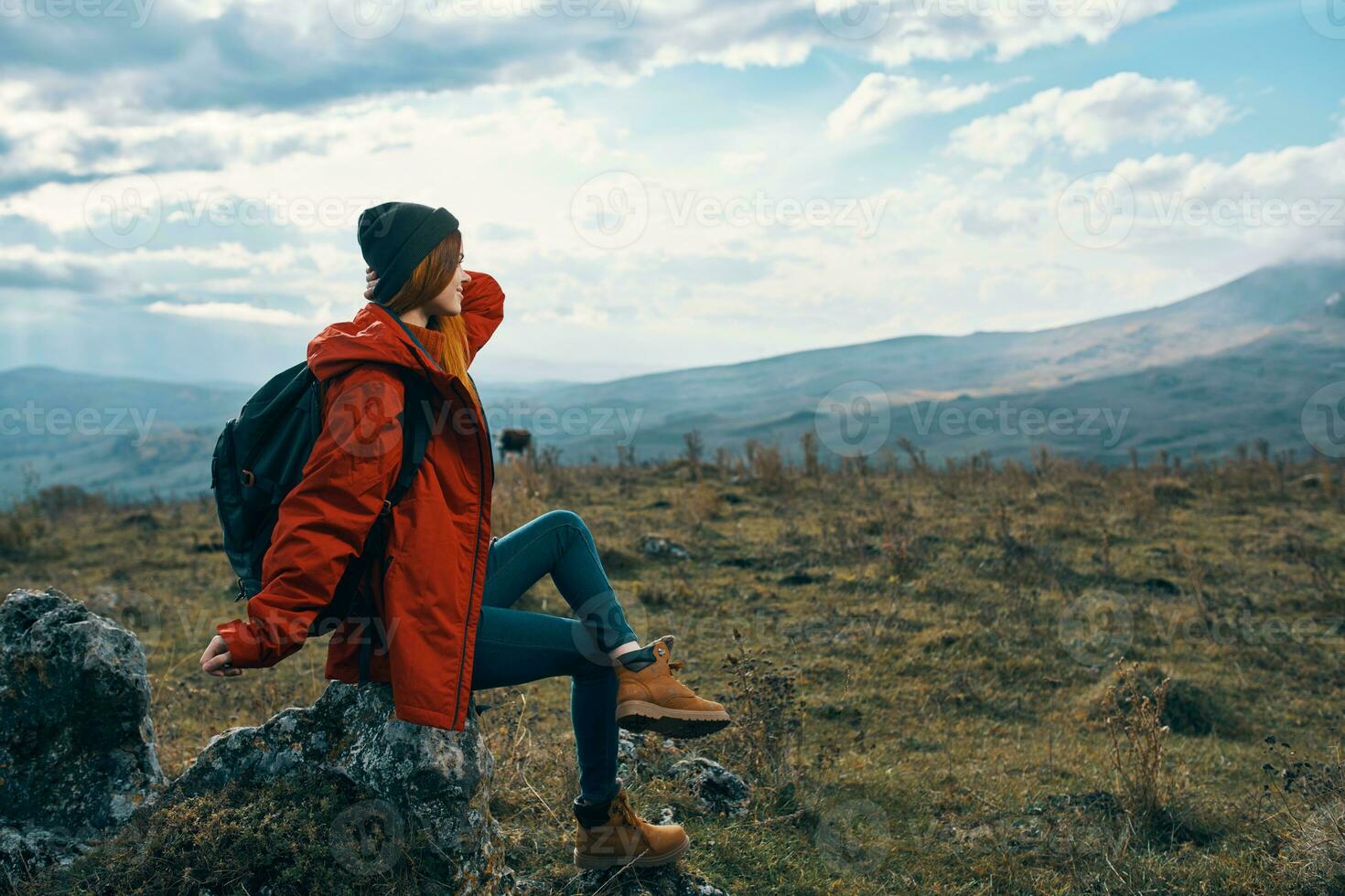 Traveler In a red jacket, hats with a backpack sits on a stone in the mountains in nature photo