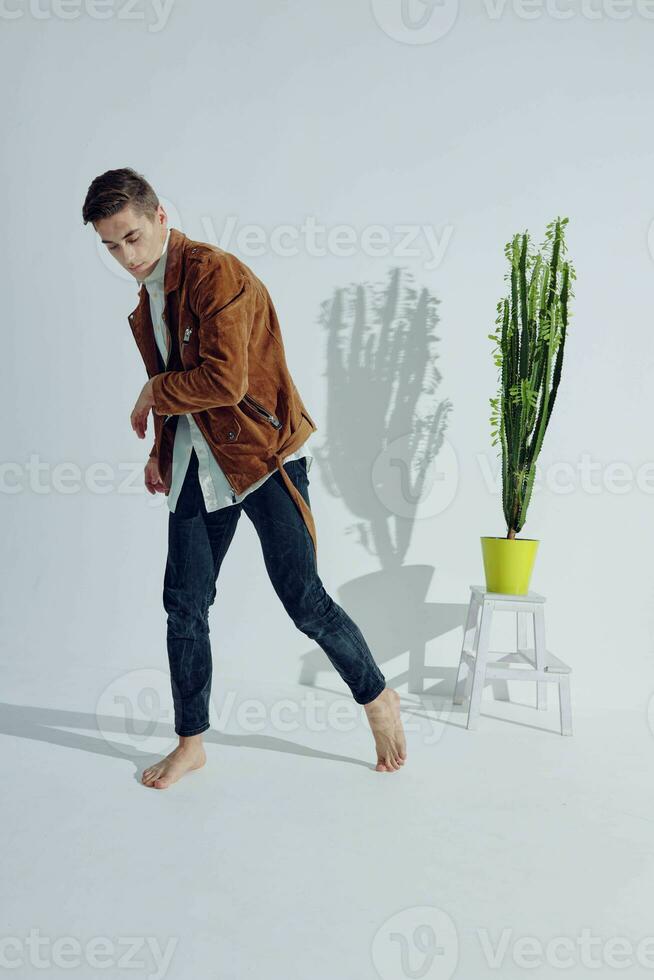 A man in a brown jacket and dark trousers stands barefoot on the floor near a flower photo