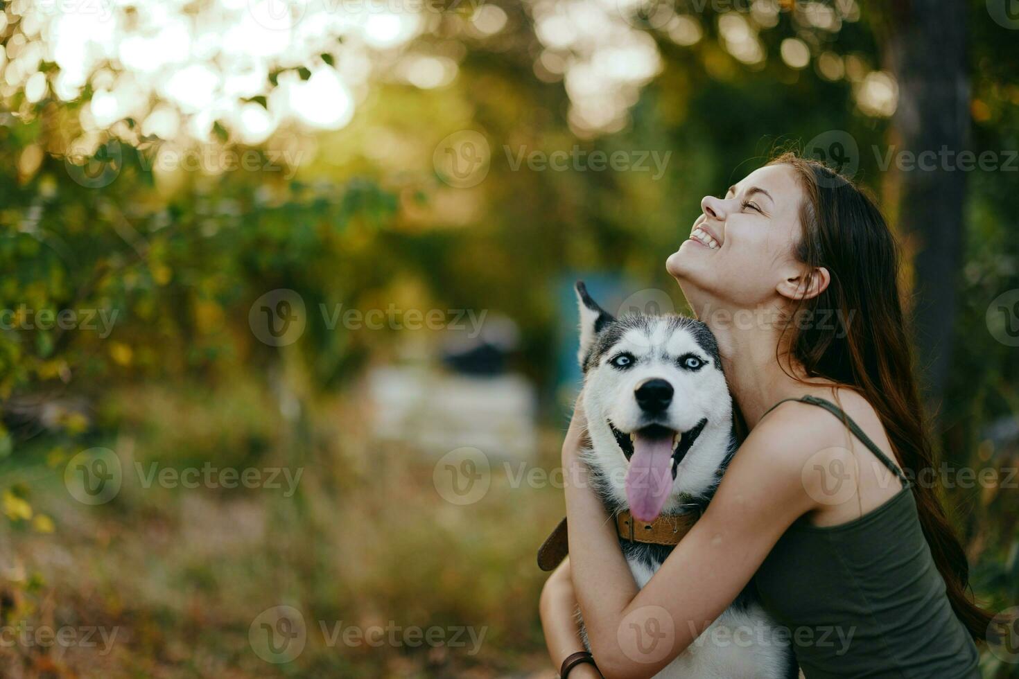 un mujer con un fornido raza perro sonrisas y afectuosamente golpes su amado perro mientras caminando en naturaleza en el parque en otoño en contra el fondo de puesta de sol foto