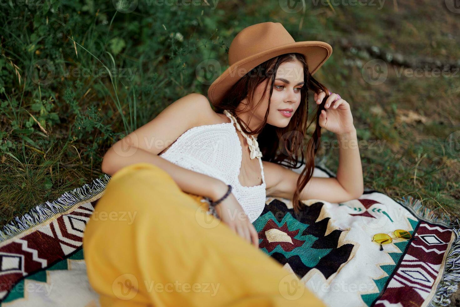 A woman in eco-friendly clothing in a hippie look is lying on a colored plaid smiling and looking at an autumn sunset in nature in the park. Lifestyle on a journey of harmony with the world photo