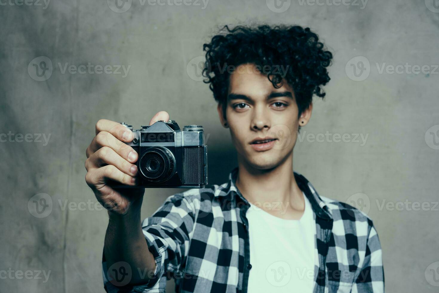 joven hombre con un cámara en mano y en un tartán camiseta en un gris antecedentes adentro fotógrafo foto