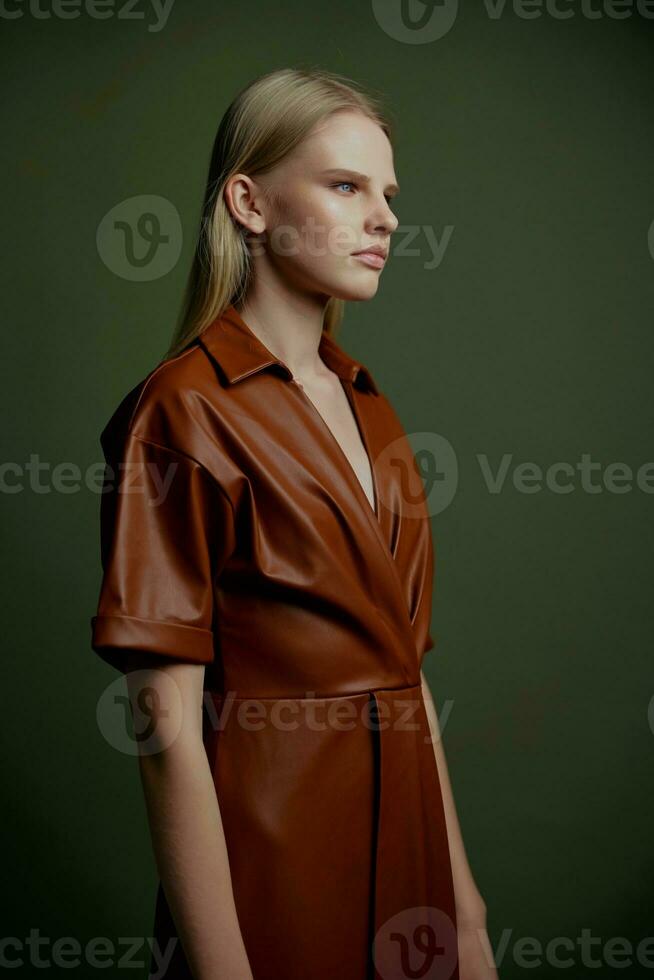 Self-confident fashionable blonde young model looking aside dressing in brown leather trench coat posing isolated on over olive green wall background. Stylish studio profile portrait offer for banner photo