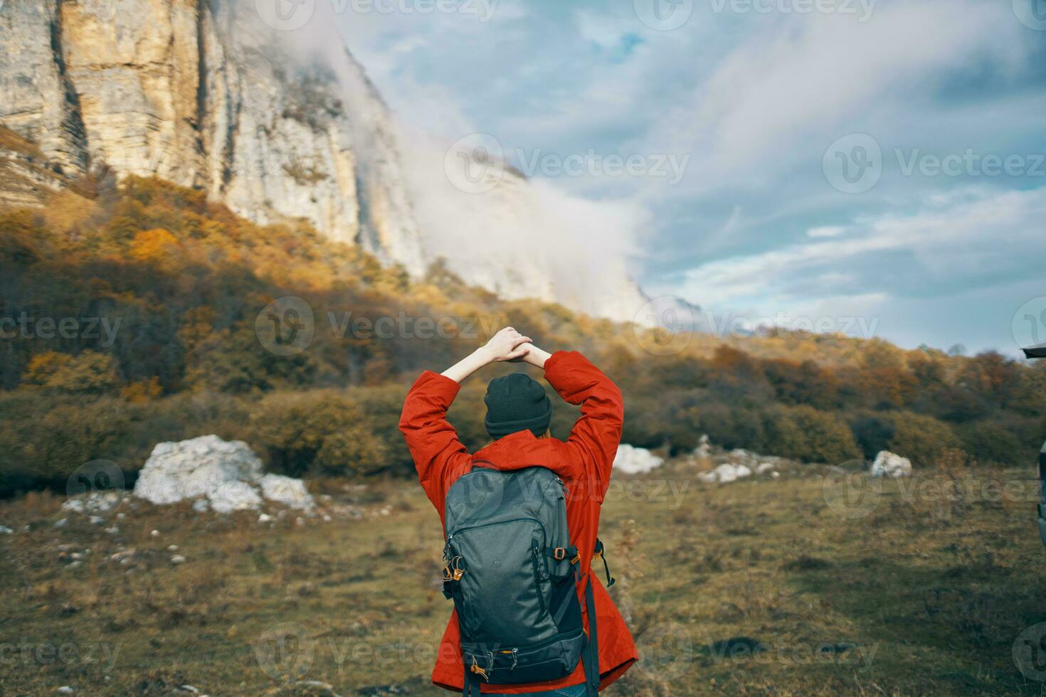 woman in a gray hat red jacket with a backpack gestures with her hands on nature in the mountains photo