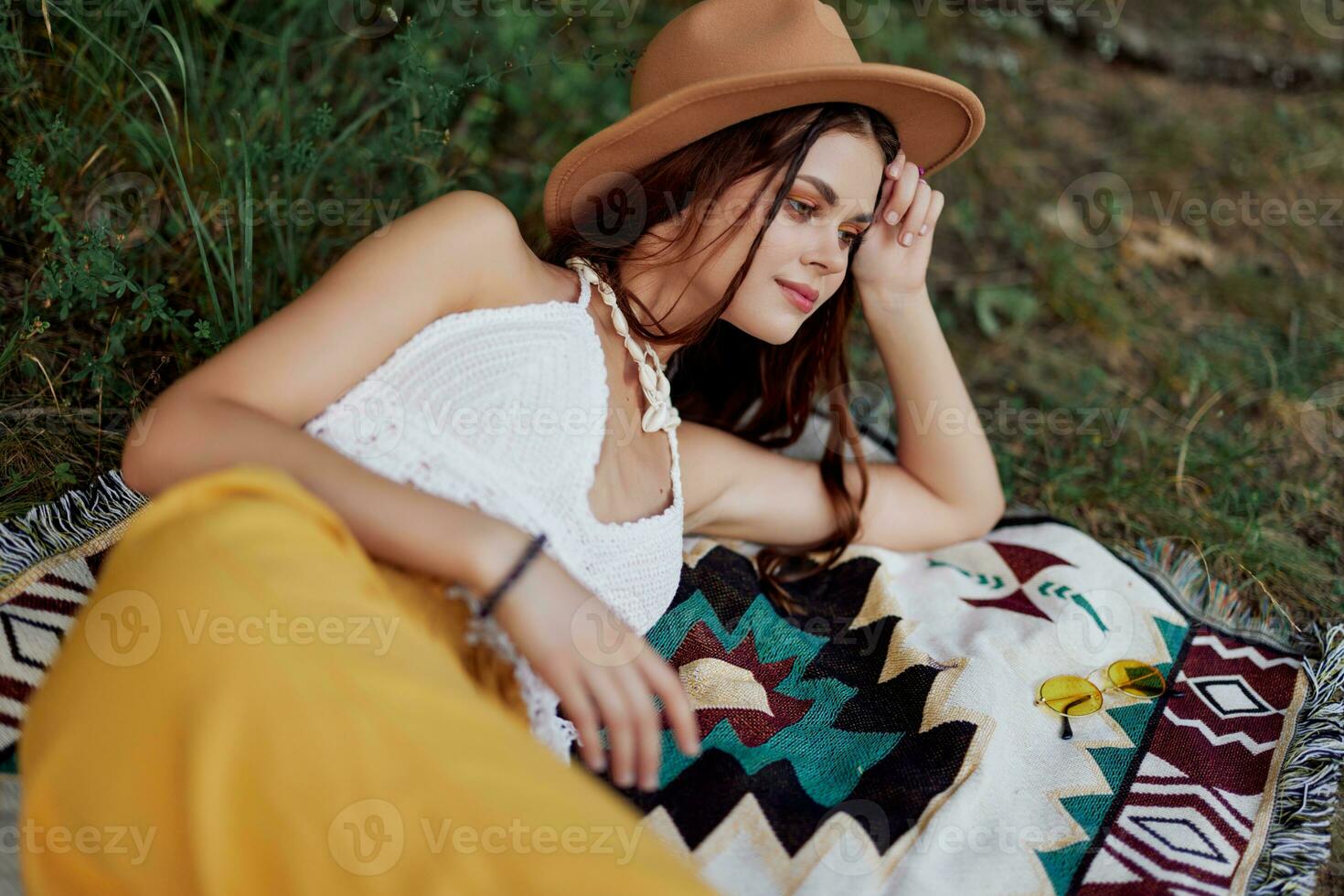 A woman in eco-friendly clothing in a hippie look is lying on a colored plaid smiling and looking at an autumn sunset in nature in the park. Lifestyle on a journey of harmony with the world photo
