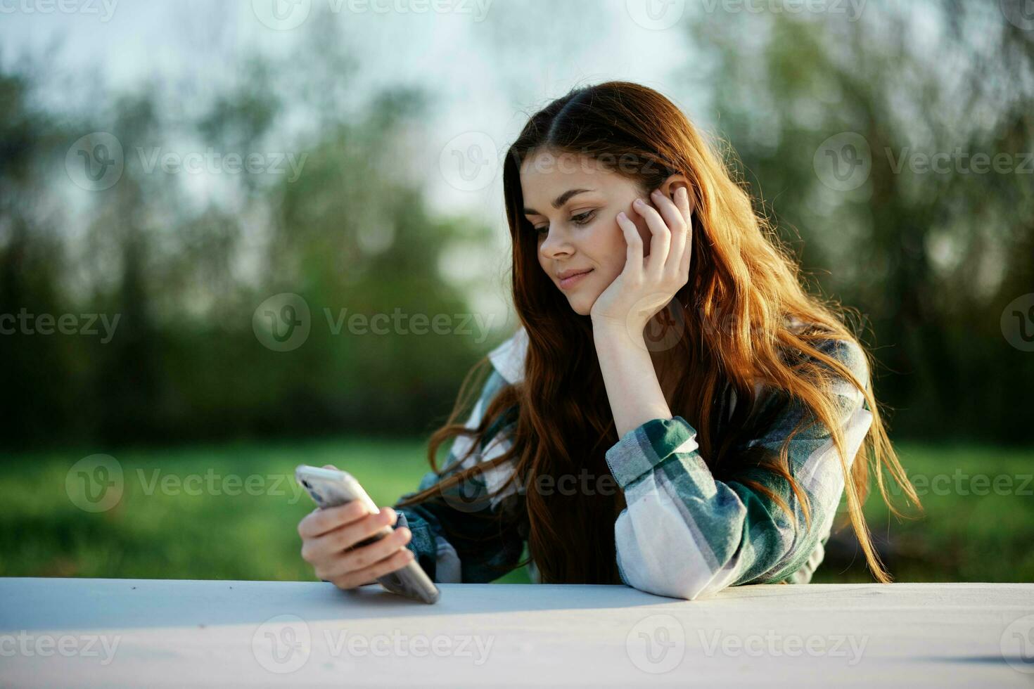 A woman with curly red hair in a plaid shirt sits outdoors in the park with a cell phone in her hands and looks away. Freelance work in the park photo