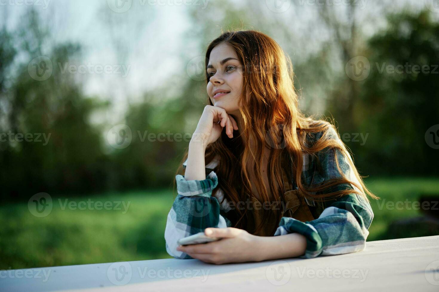 Girl with phone in hand sitting in nature in the park at the table smiling beautifully and looking at the camera with her red hair lit by the sunset sunlight of summer photo