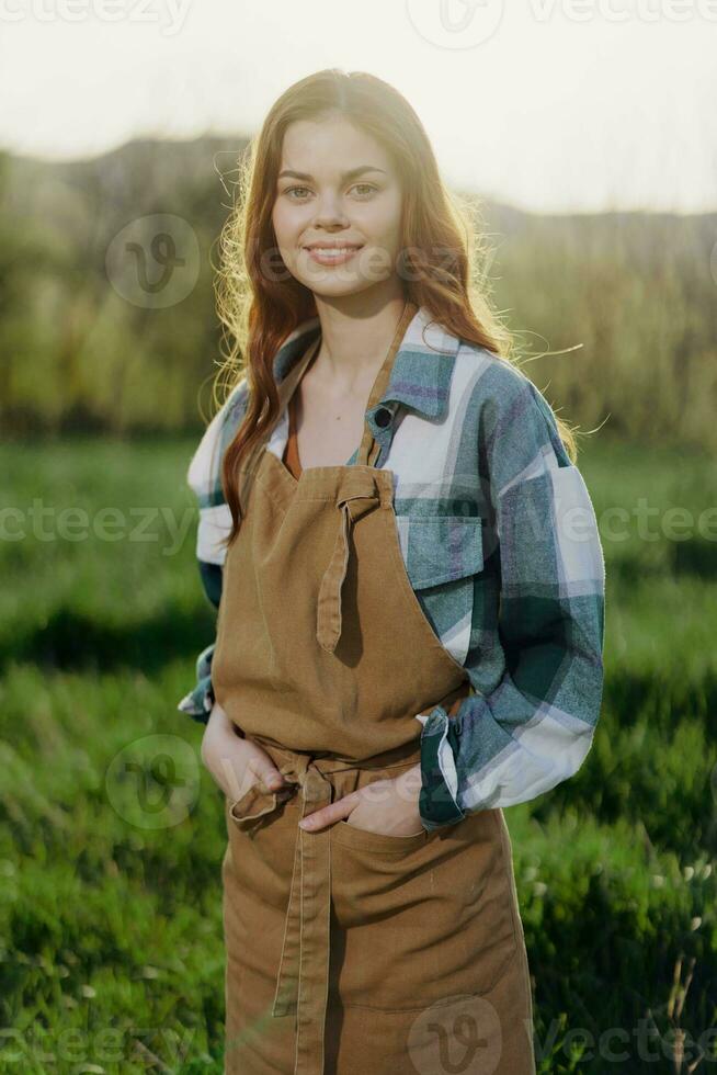 A woman gardener in an apron stands in a field of green grass outdoors, smiling on a summer day into a sunset photo