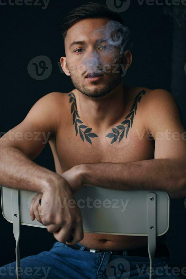 Modelling snapshots. Pensive frustrated tanned attractive handsome naked man sit on chair smoking looks at camera posing isolated in black studio background. Fashion offer. Copy space for ad. Closeup photo