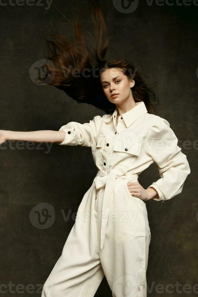 a pretty brunette in a light jumpsuit gestures with her hands on a dark background photo