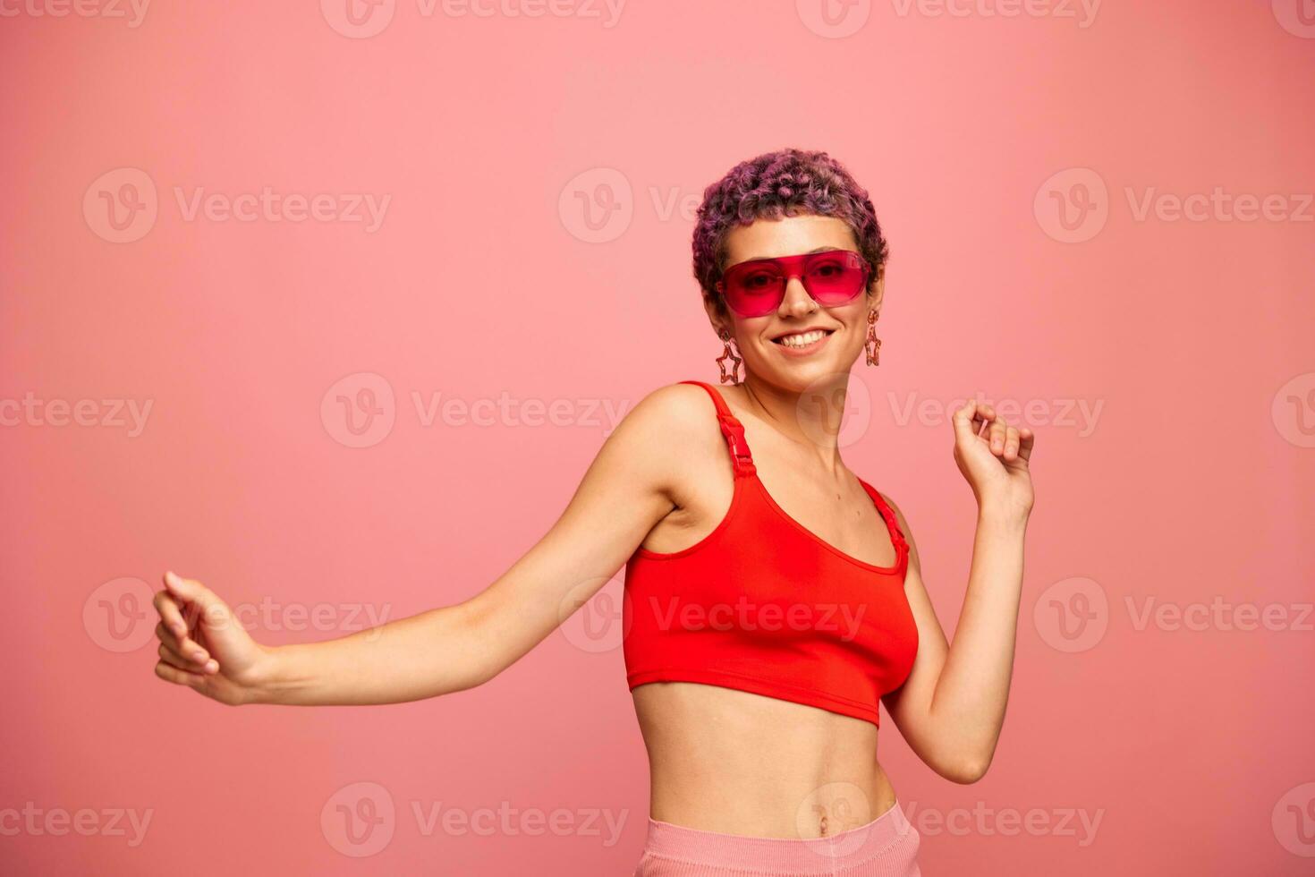 Fashion portrait of a woman with a short haircut in colored sunglasses with unusual accessories with earrings smiling on a pink bright background with a fitness body dancing photo