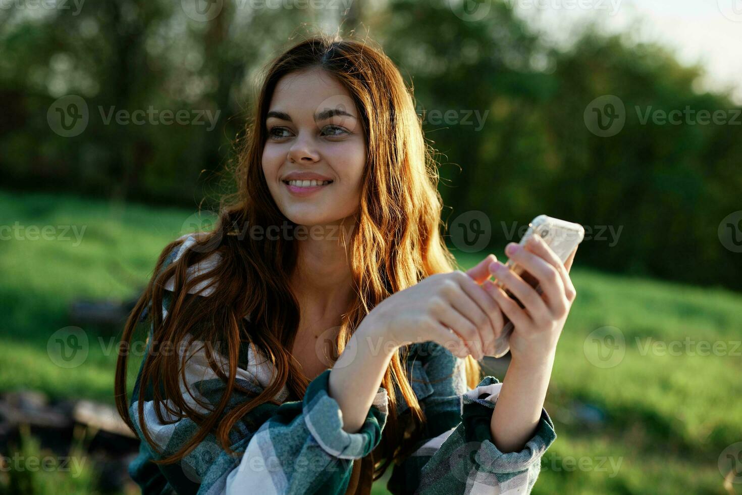 un hermosa mujer relajante y trabajando en su teléfono sentado en naturaleza en el parque entre el arboles sonriente y participación su teléfono inteligente en su mano iluminado por el brillante puesta de sol ligero foto