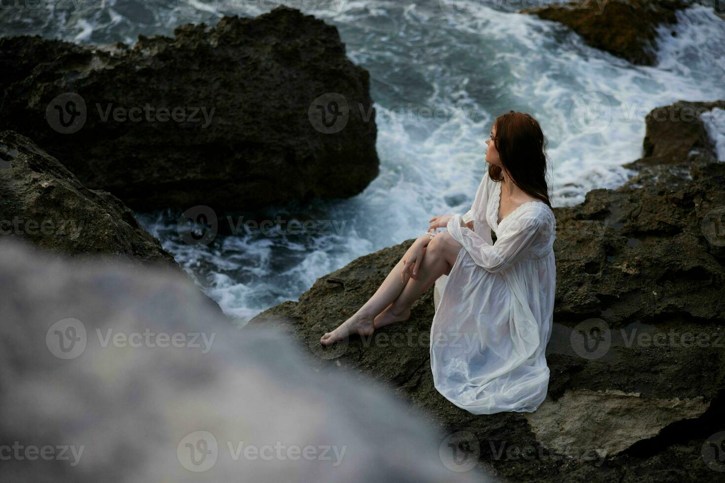 un mujer en un blanco vestir descalzo se sienta en un Roca en un acantilado parte superior ver foto