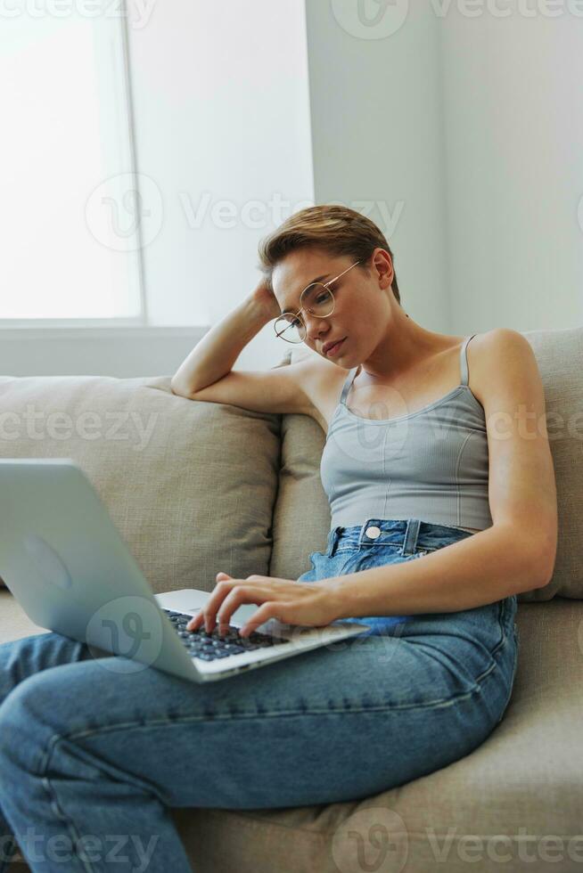 Teenage girl freelancer with laptop sitting on couch at home smiling in home clothes and glasses with short haircut, lifestyle with no filters, free copy space photo