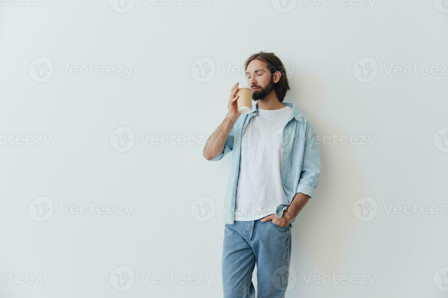 Lanza libre milenario hombre con un barba Bebiendo café desde un reciclado taza en elegante hipster ropa blanco camiseta azul pantalones y camisa en un blanco antecedentes foto